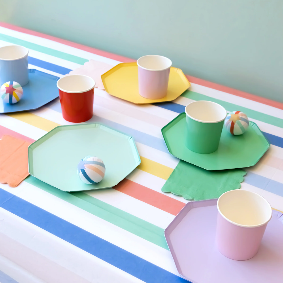 A colorful party table setup featuring octagonal paper plates, matching cups, napkins, and surprise balls arranged on a pastel striped tablecloth.