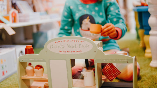 Wooden toy cafe stand reading "Bird's Nest Cafe, baked fresh and sweet, served all day long" as a child in a green polka dot jumper sips tea