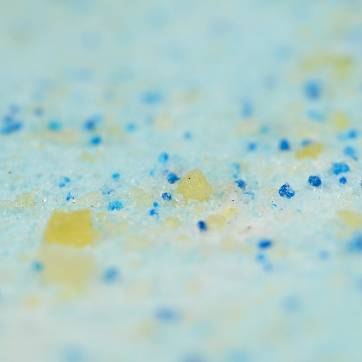 Close-up of a granular mixture of blue and yellow particles on a light blue background, resembling bath salt. The texture is coarse and colorful.