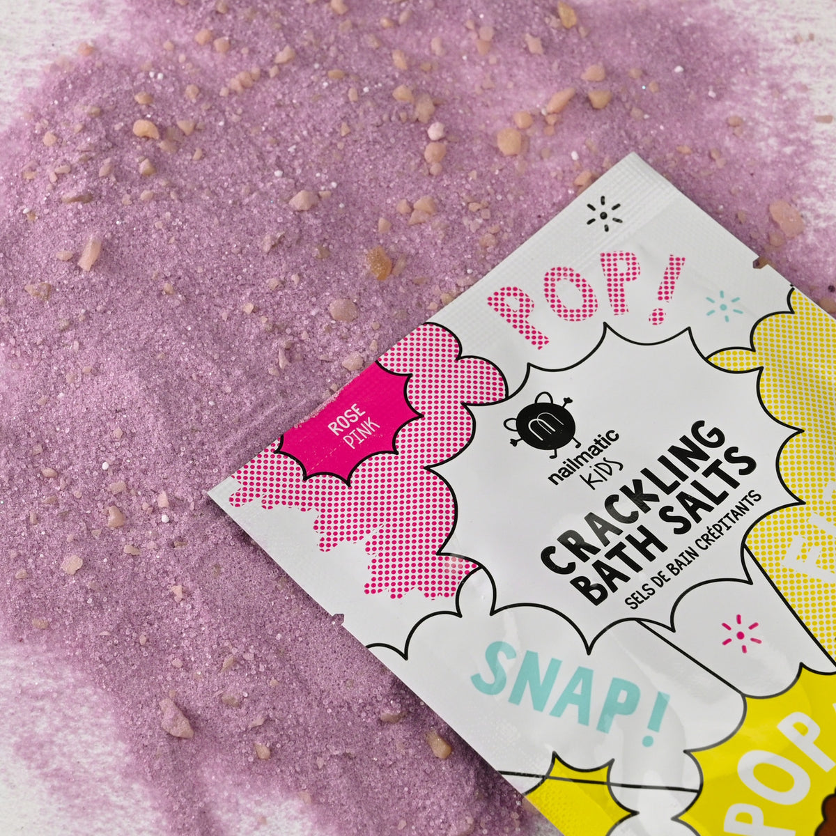 Packet of crackling bath salts labeled "Pop!" and "Snap!" on a playful design. Rose pink salts are scattered underneath, adding a whimsical, fun vibe.
