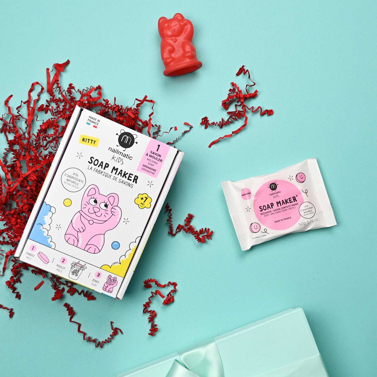 Colorful DIY soap kit for kids with cartoon cat illustrations on the box. Includes a red cat soap mold, white soap packet, and red crinkle paper on a turquoise background. Playful and creative tone.