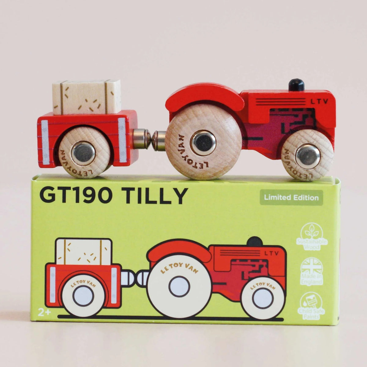 Red wooden toy tractor with trailer resting on top of its green packaging box in a side view.