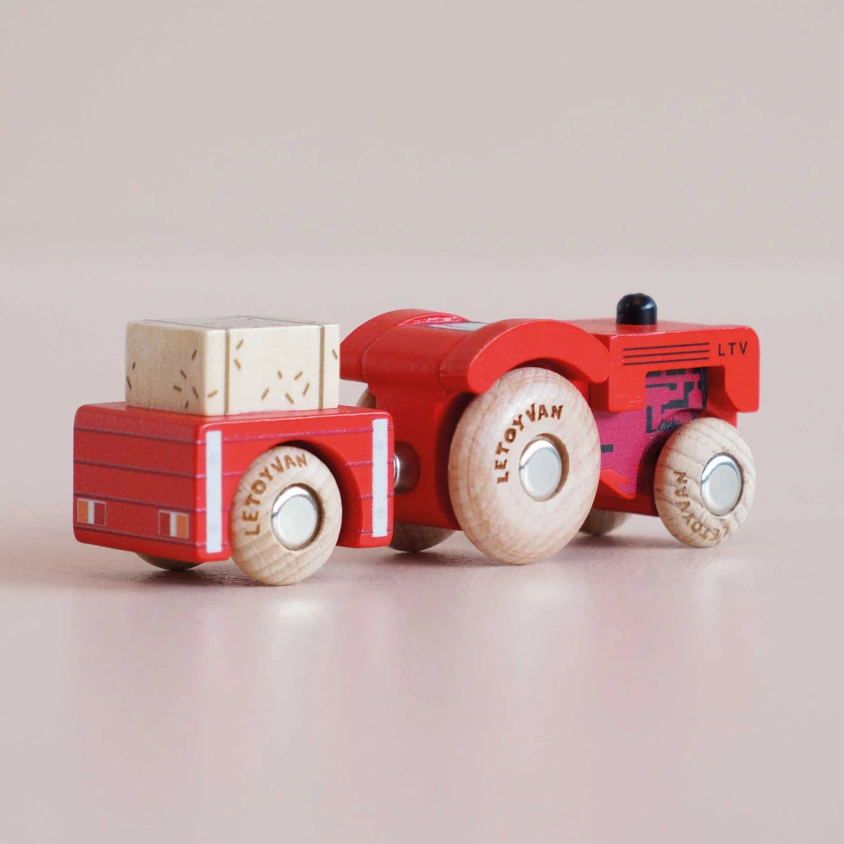 Red wooden toy tractor with trailer carrying a hay bale, captured in a rear three-quarter view.