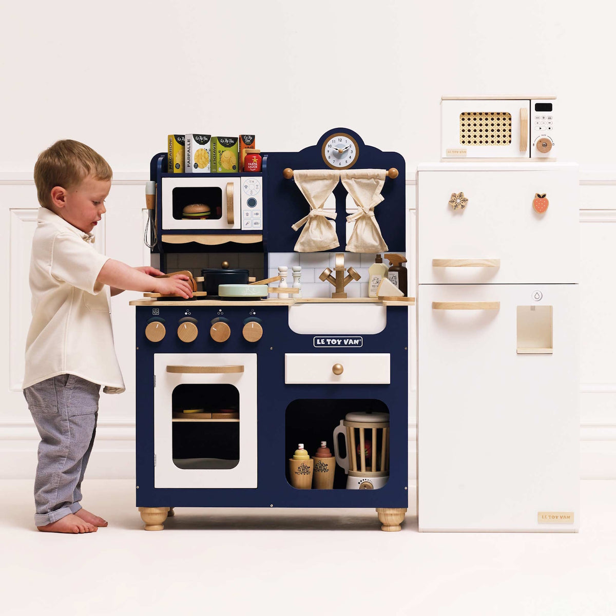 A toddler playing with a blue wooden toy kitchen set next to a white toy fridge.