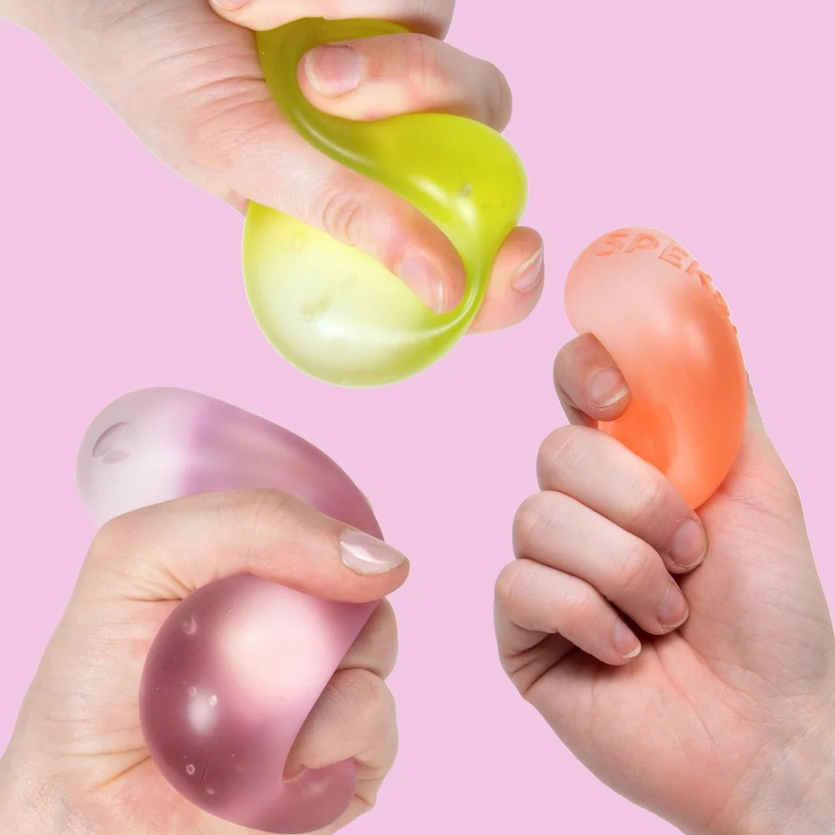 Three hands squeezing and squishing colorful memory gel stress balls on a pink background.
