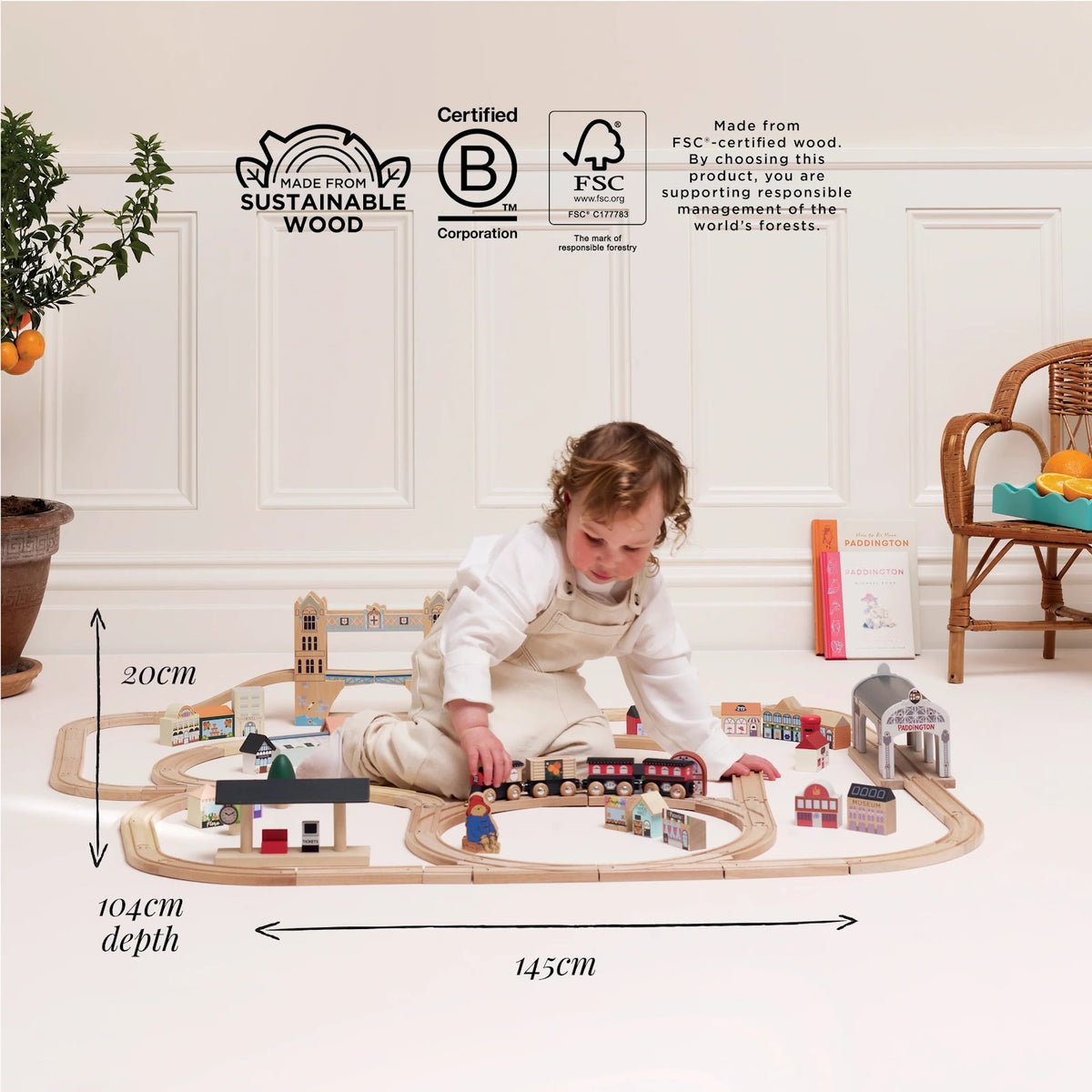 A child playing on the floor with a wooden train set, featuring overlaid dimensions and sustainability logos.