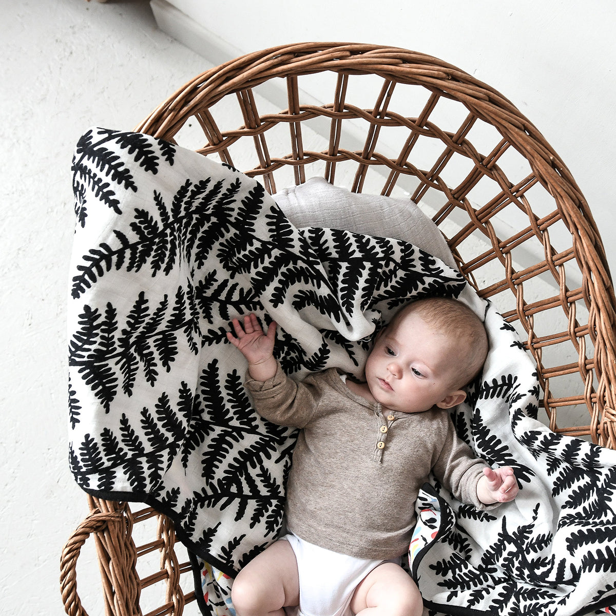 Baby relaxing in a wicker chair on a black-and-white fern printed muslin blanket.