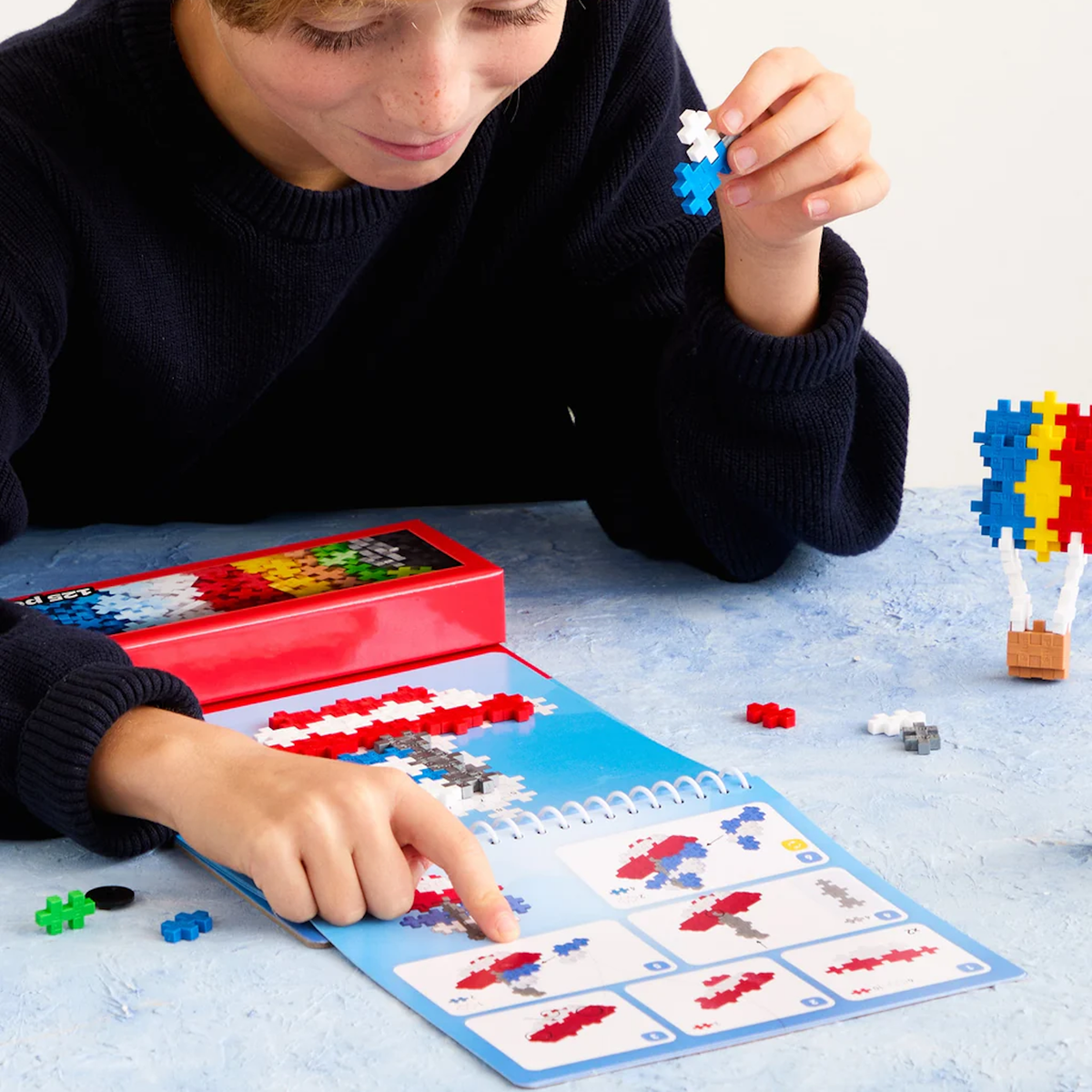 Child follows instructions on a Plus-Plus activity pad to build a hot air balloon with colorful building blocks.