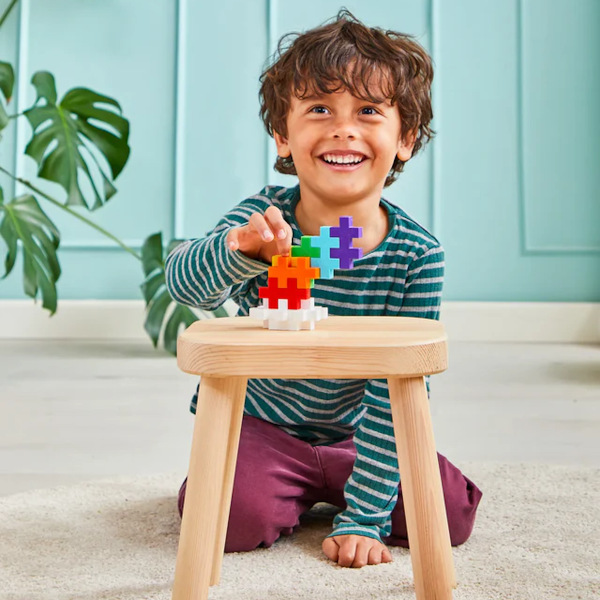 Smiling child with curly hair, wearing a striped shirt, builds with colorful plus-shaped blocks on a wooden stool. A large plant decorates the background.