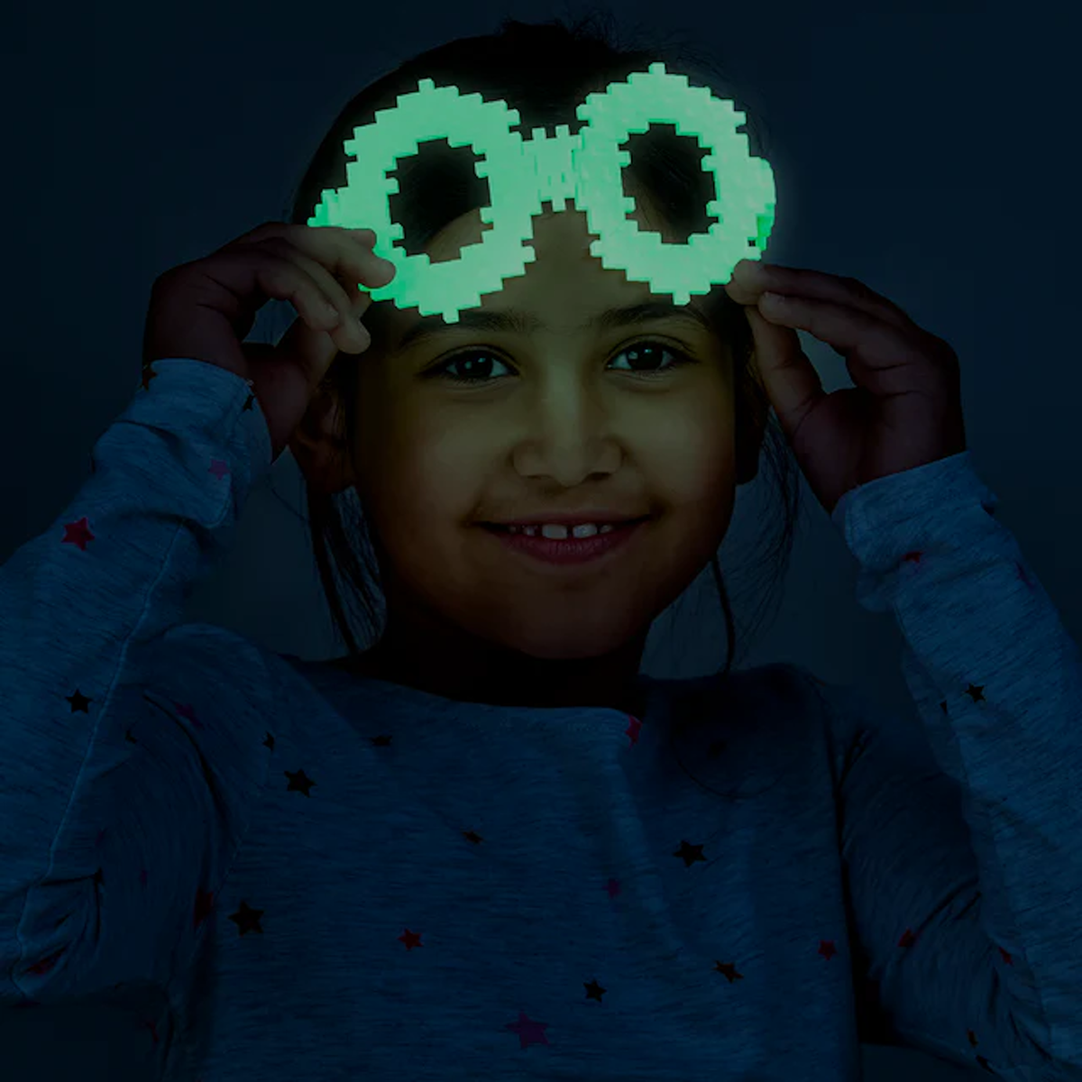A smiling child wearing pixelated, glowing green glasses holds them up with both hands in a dark setting, creating a playful and fun atmosphere.
