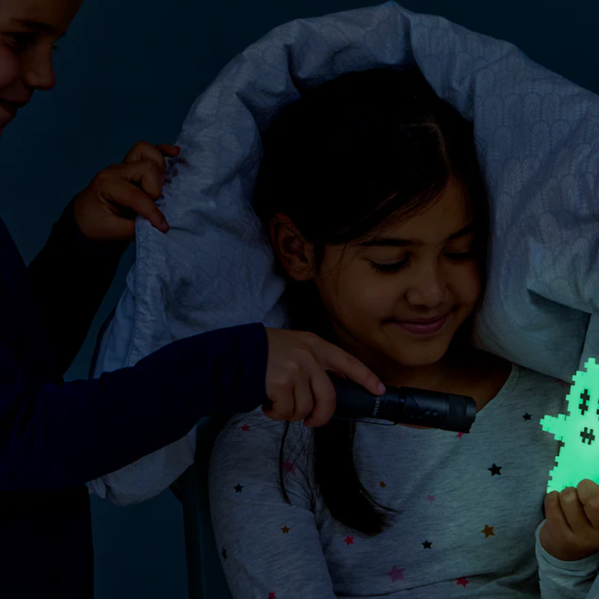 Two children are under a blanket, one holding a glowing pixelated ghost toy. They appear excited and playful, enjoying a cozy night adventure.