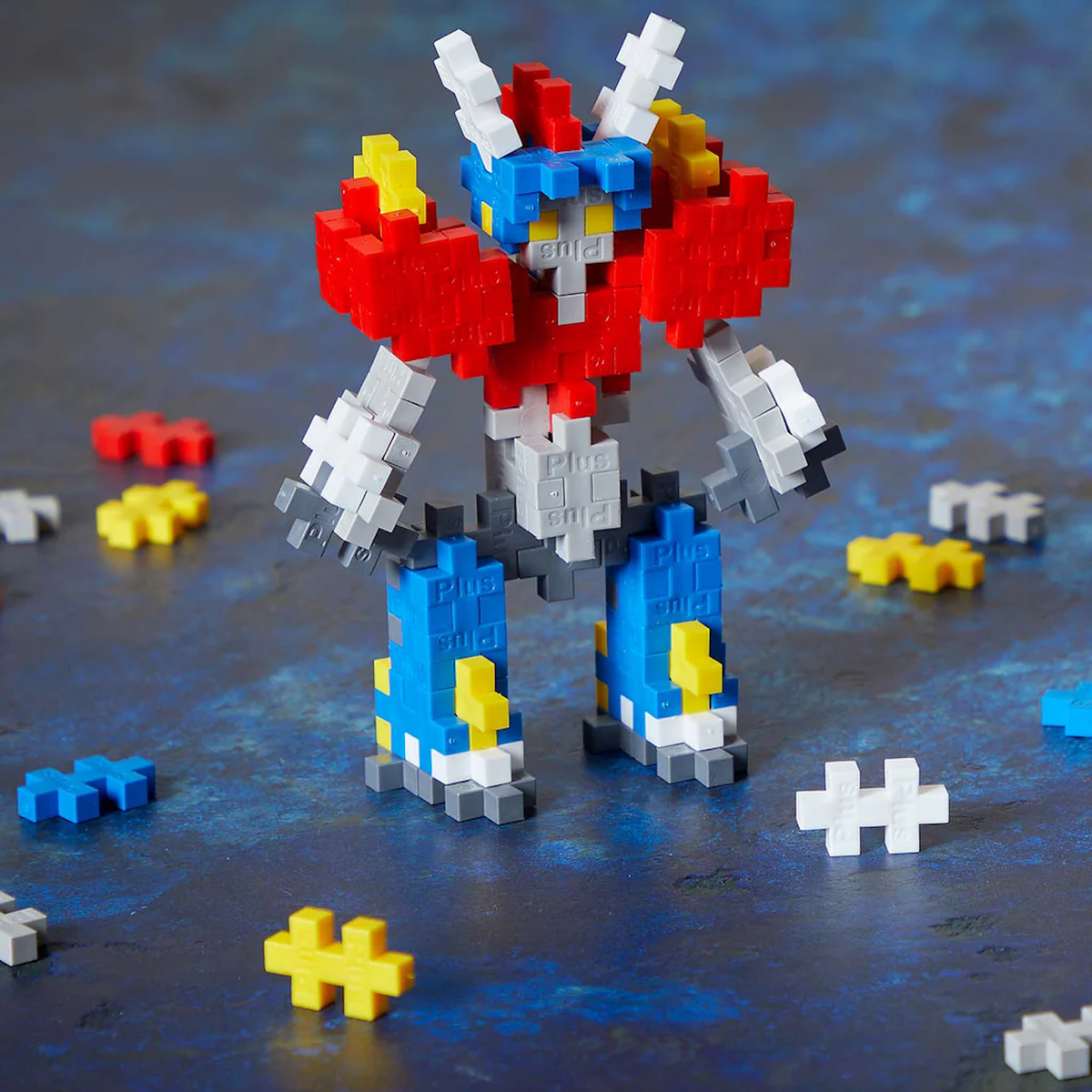 Colorful mecha robot figure made from red, blue, and white Plus-Plus building blocks on a blue textured surface.