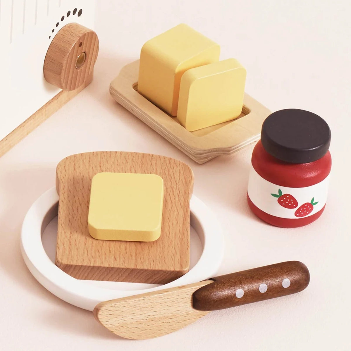 Wooden toy breakfast accessories including a butter dish, toast on a plate, strawberry jam, and a knife.