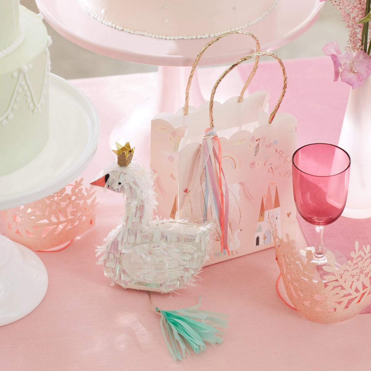 Pink party table with a swan piñata favors bag, small crowns, cakes, and a pink glass.