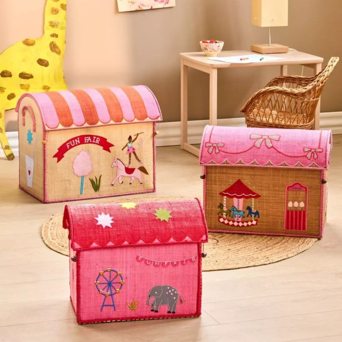 Three pink and tan raffia toy baskets shaped like houses with a funfair theme on a wooden floor.