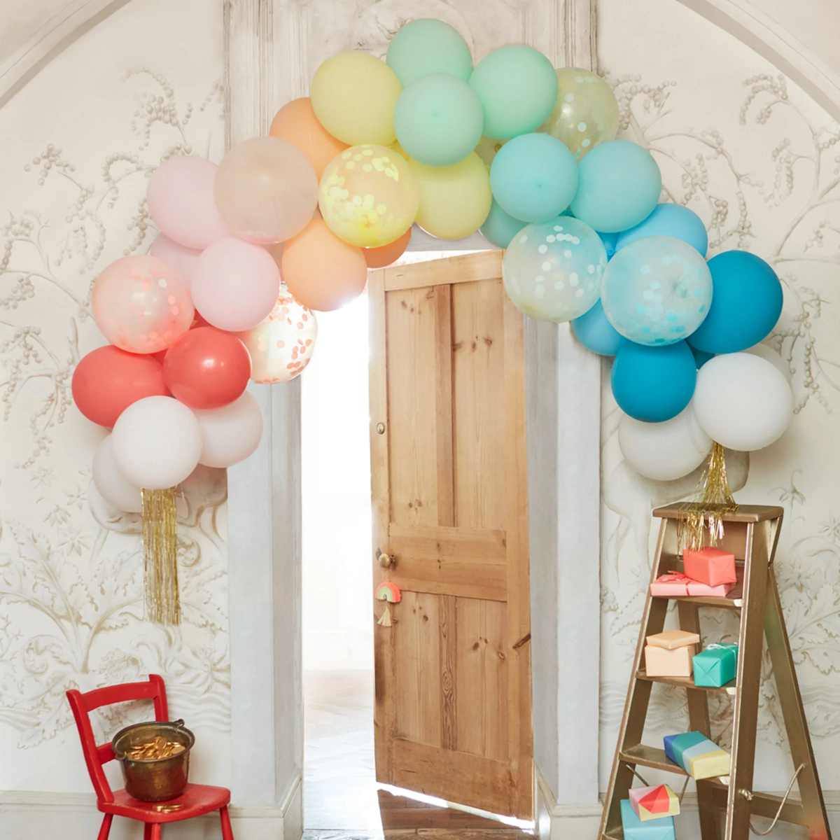 Large rainbow balloon arch with gold tassels displayed over a wooden doorway in a decorated party setting.