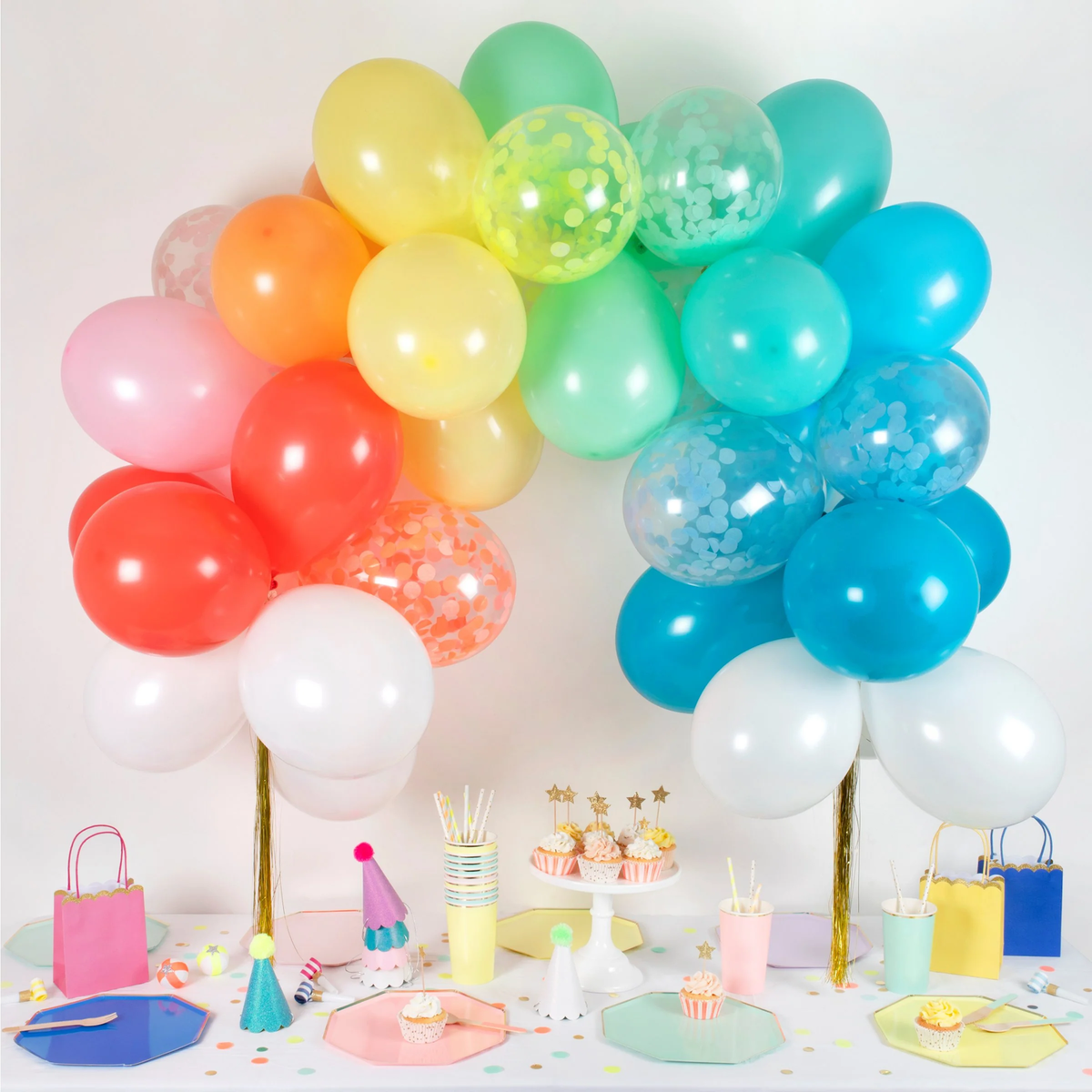 Rainbow balloon arch with gold tassels positioned over a colorful party table set with party tableware and treats.