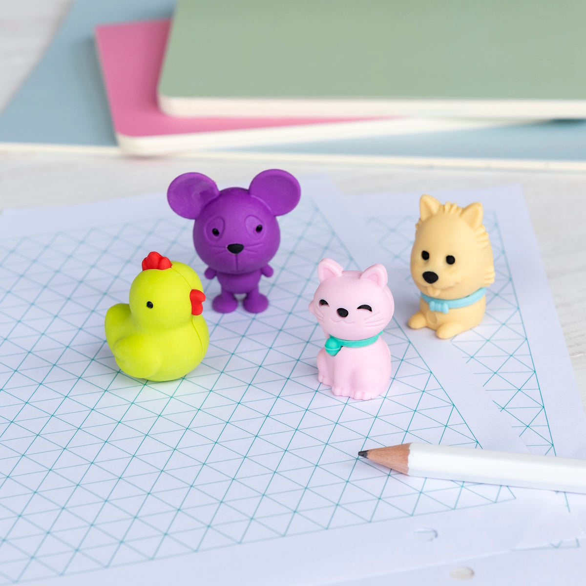 Colorful animal erasers, including a yellow duck, purple mouse, pink cat, and beige dog, on grid paper near pastel notebooks and a pencil. Playful tone.