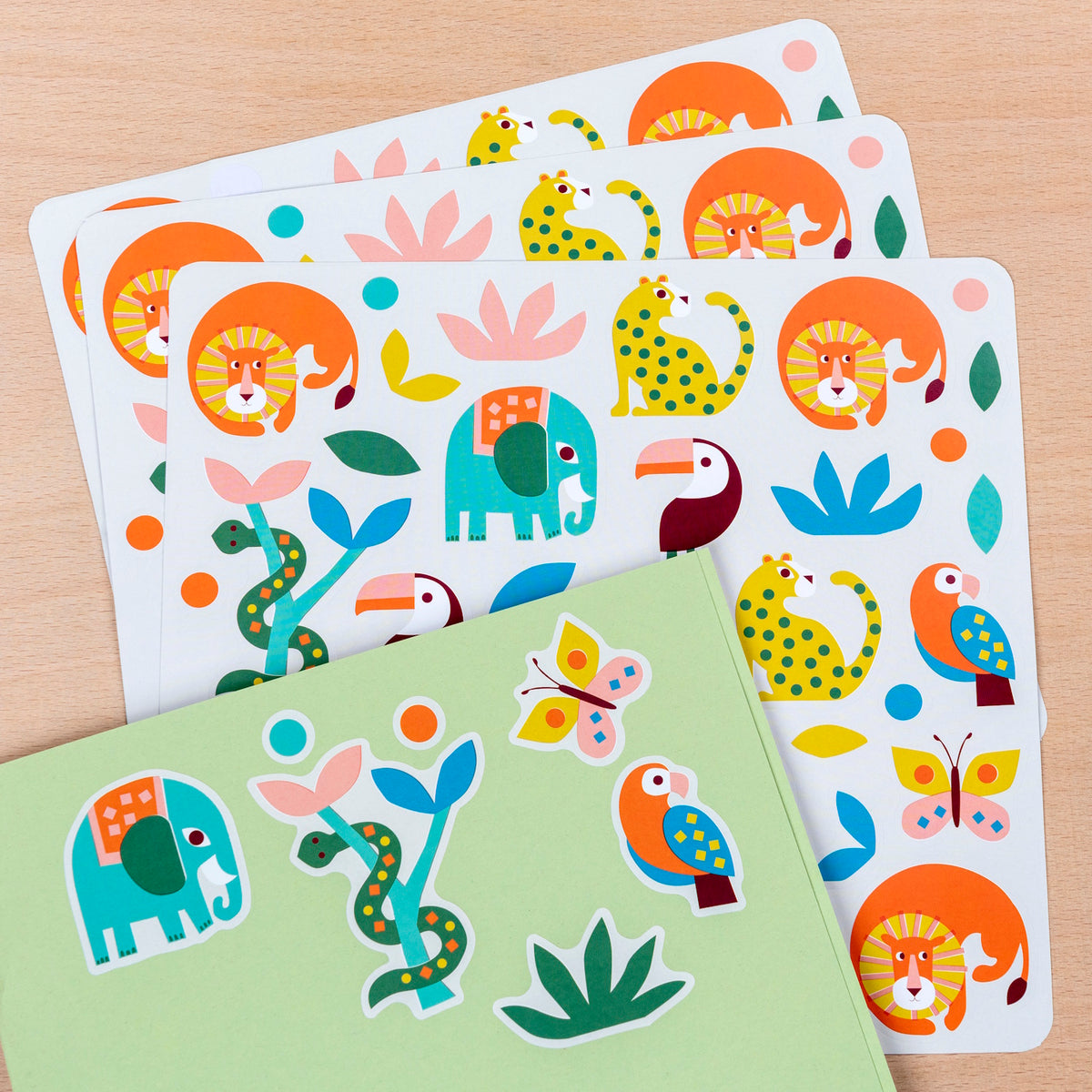 Colorful sticker sheets with playful jungle animals like elephants, lions, toucans, and butterflies on a light wooden surface, conveying a fun and lively tone.