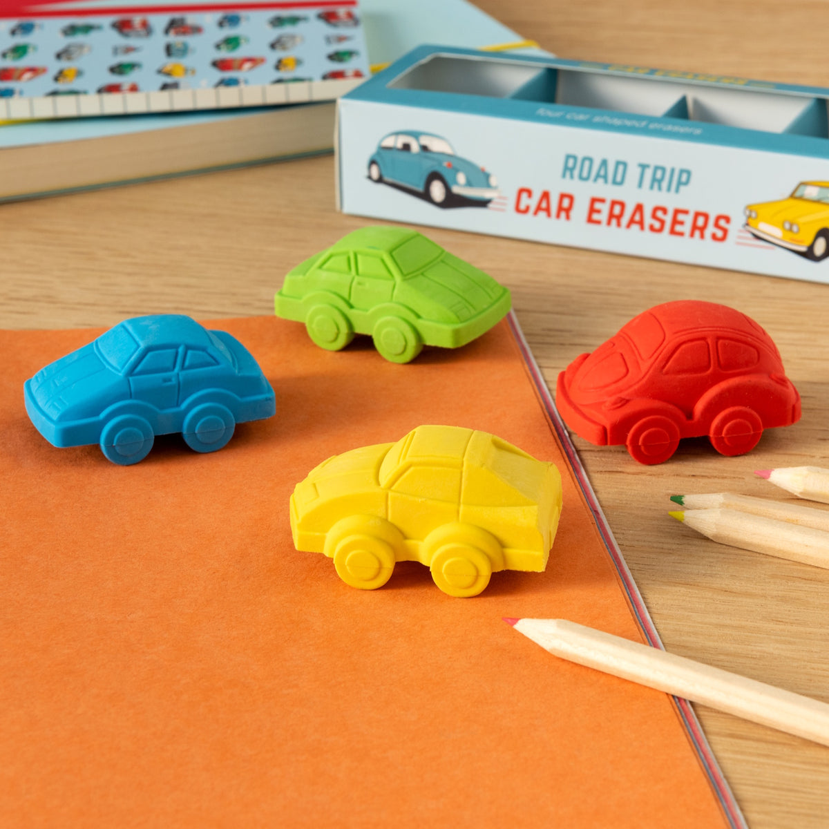 Colorful car-shaped erasers in blue, green, red, and yellow are arranged on an orange notebook. Nearby are colored pencils and a car-themed box.