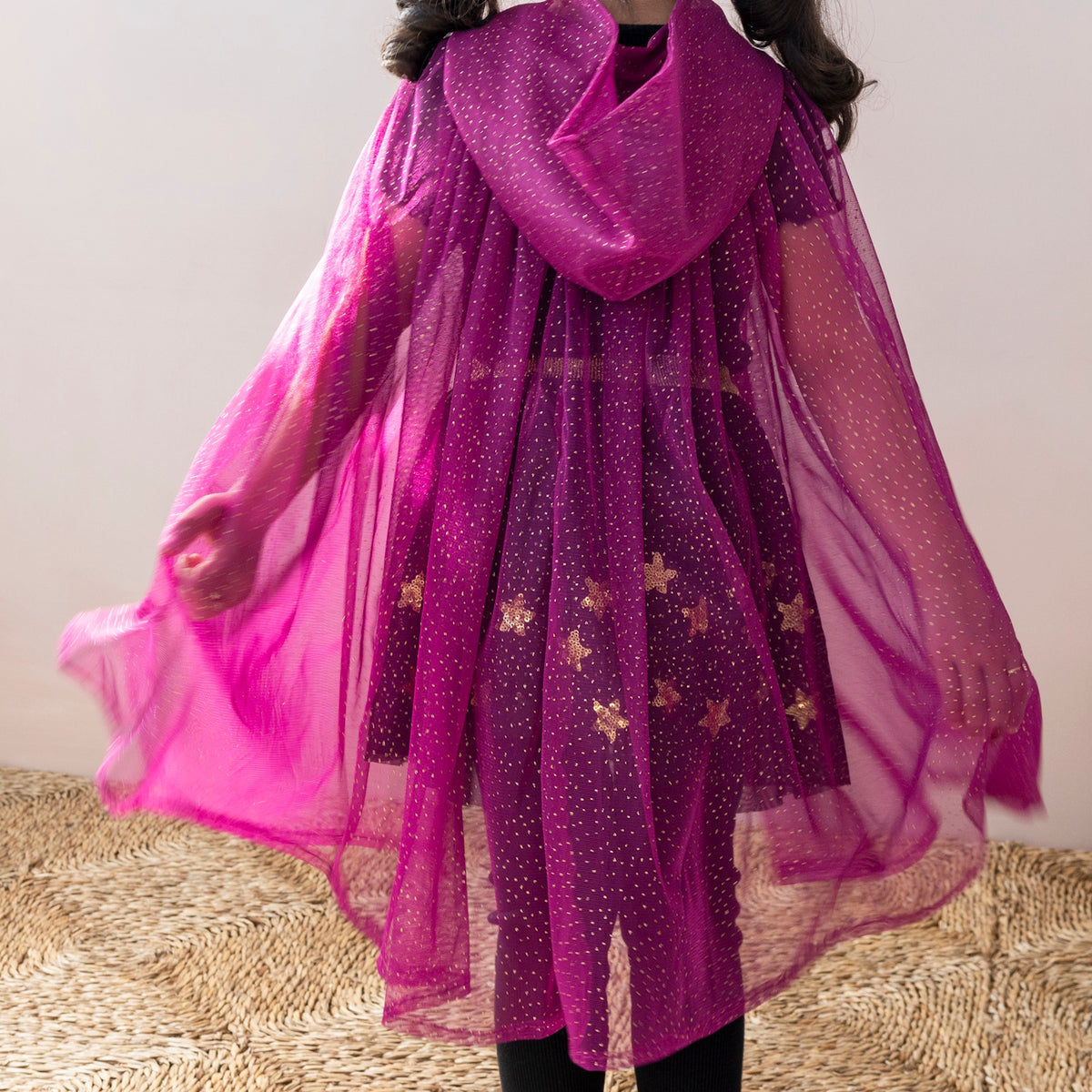 A child is seen from behind wearing a vibrant, shimmering purple cape with golden star patterns. The setting is indoors on a woven carpet.