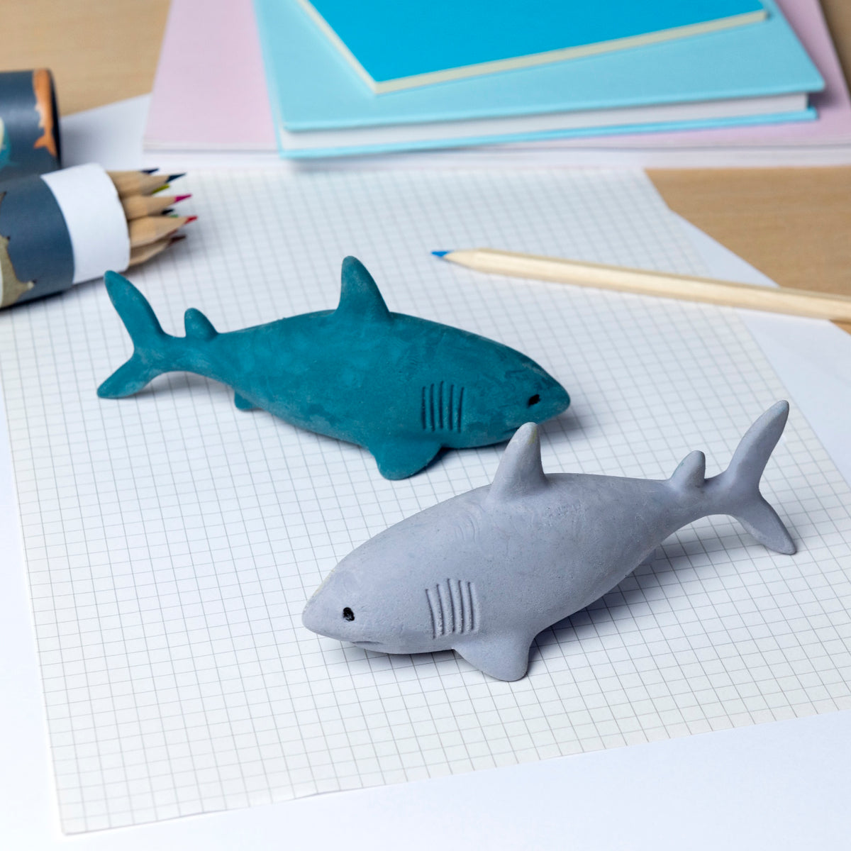 Two shark-shaped erasers, one gray and one teal, lie on graph paper. Colored pencils and pastel notebooks are nearby, creating a playful stationery scene.
