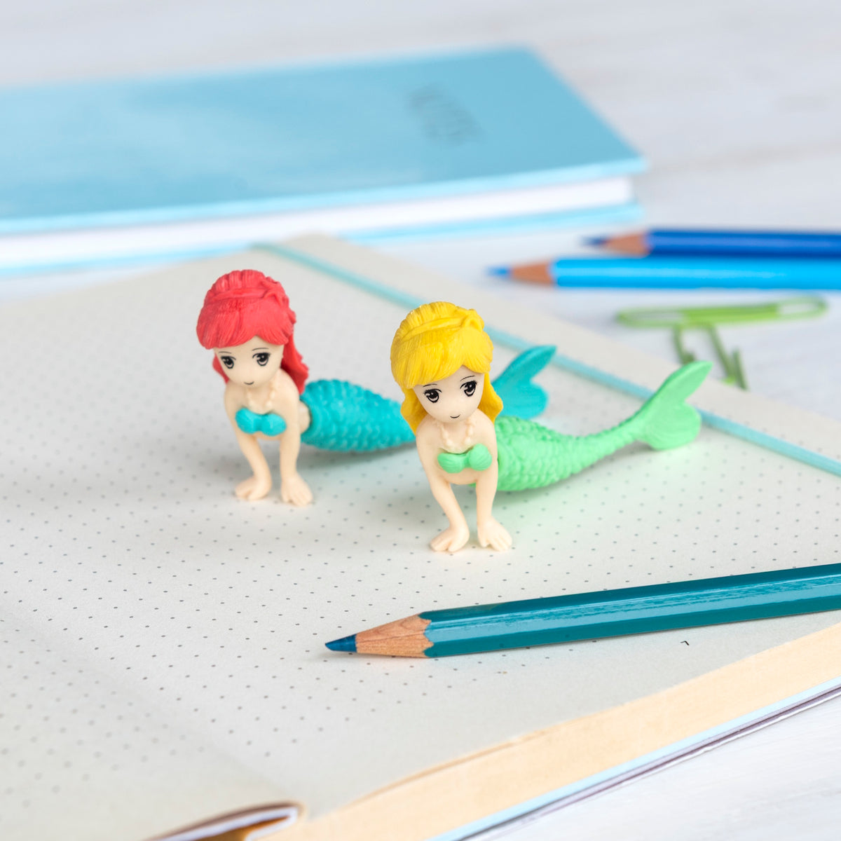 Two small mermaid figurines with colorful tails and hair on a dotted notebook page, surrounded by blue and teal pencils. Playful and creative mood.