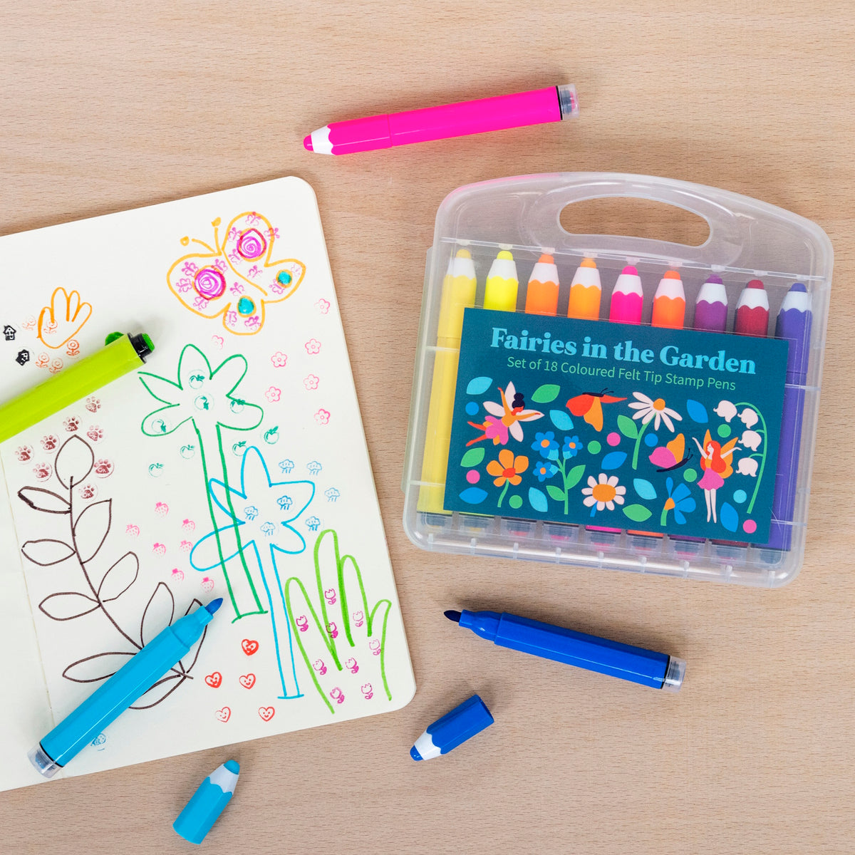 A colorful set of 18 felt tip pens labeled "Fairies in the Garden" sits beside an open sketchbook filled with doodles of flowers and a butterfly. A vibrant and creative atmosphere is conveyed.