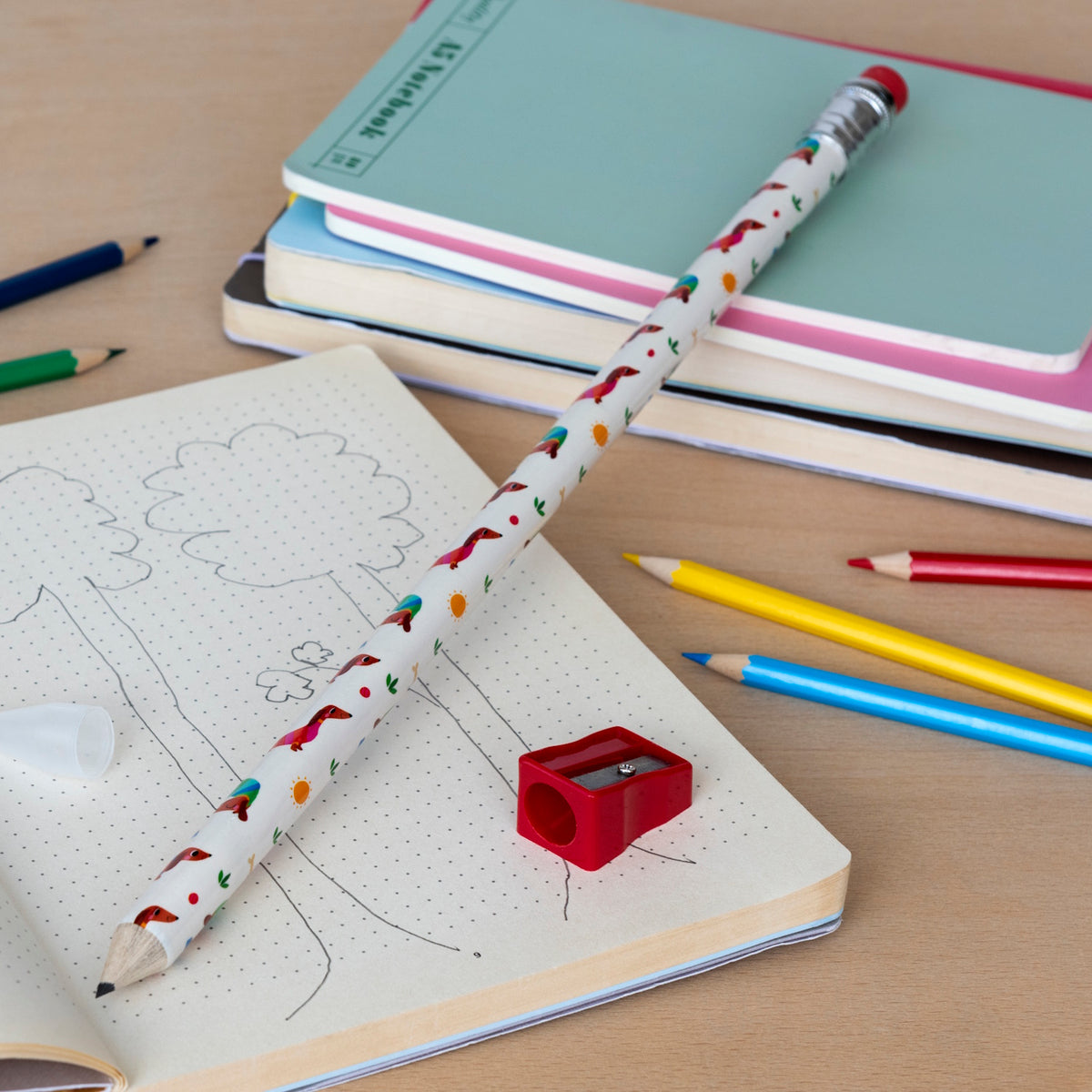 A whimsical scene with a large, printed pencil and red sharpener on a dotted notebook featuring tree sketches. Colored pencils and pastel notebooks add a playful touch.