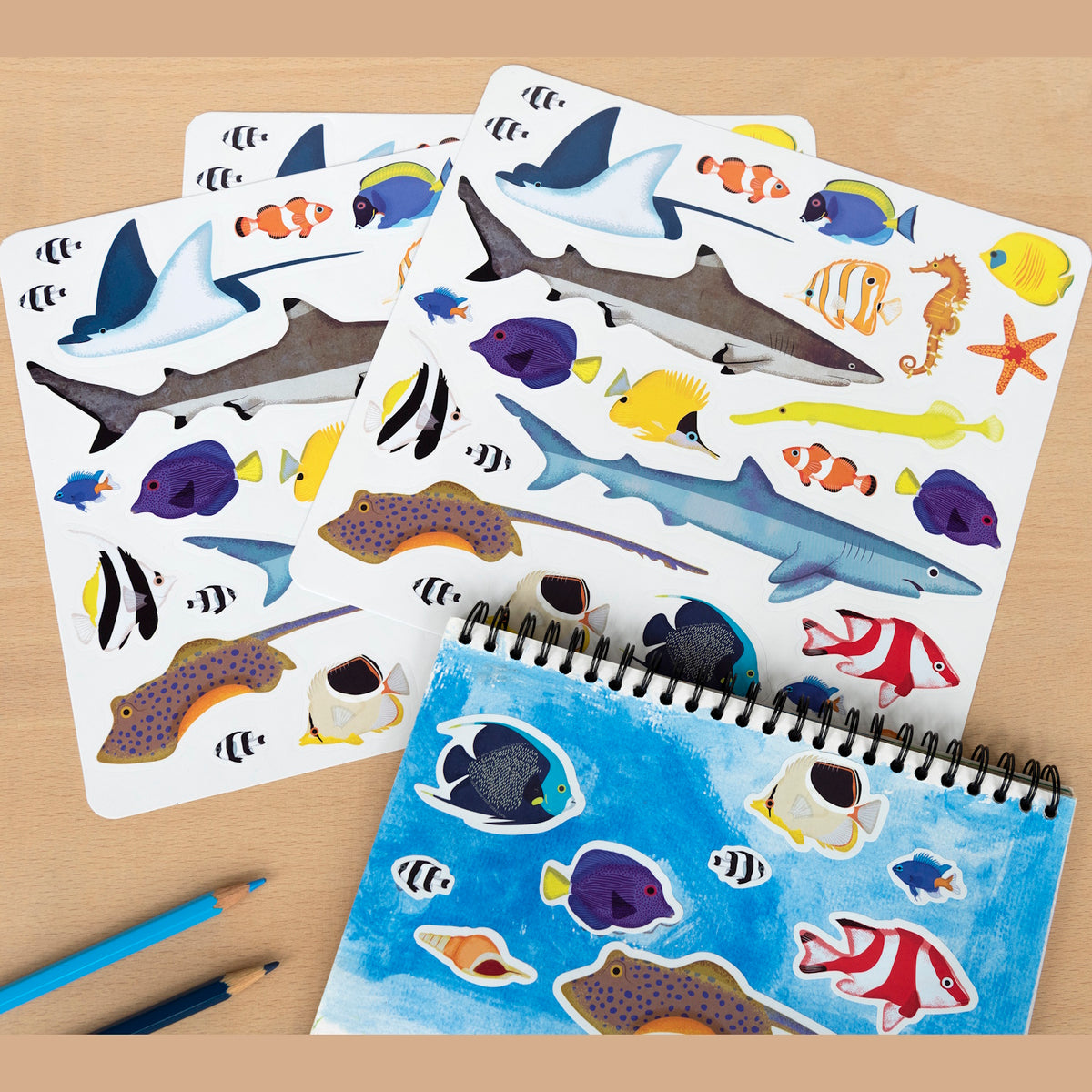 Sticker sheets featuring vibrant marine life, like sharks, rays, seahorses, and colorful fish, lie beside a sketchbook with similar ocean stickers. Two blue pencils complement the scene.