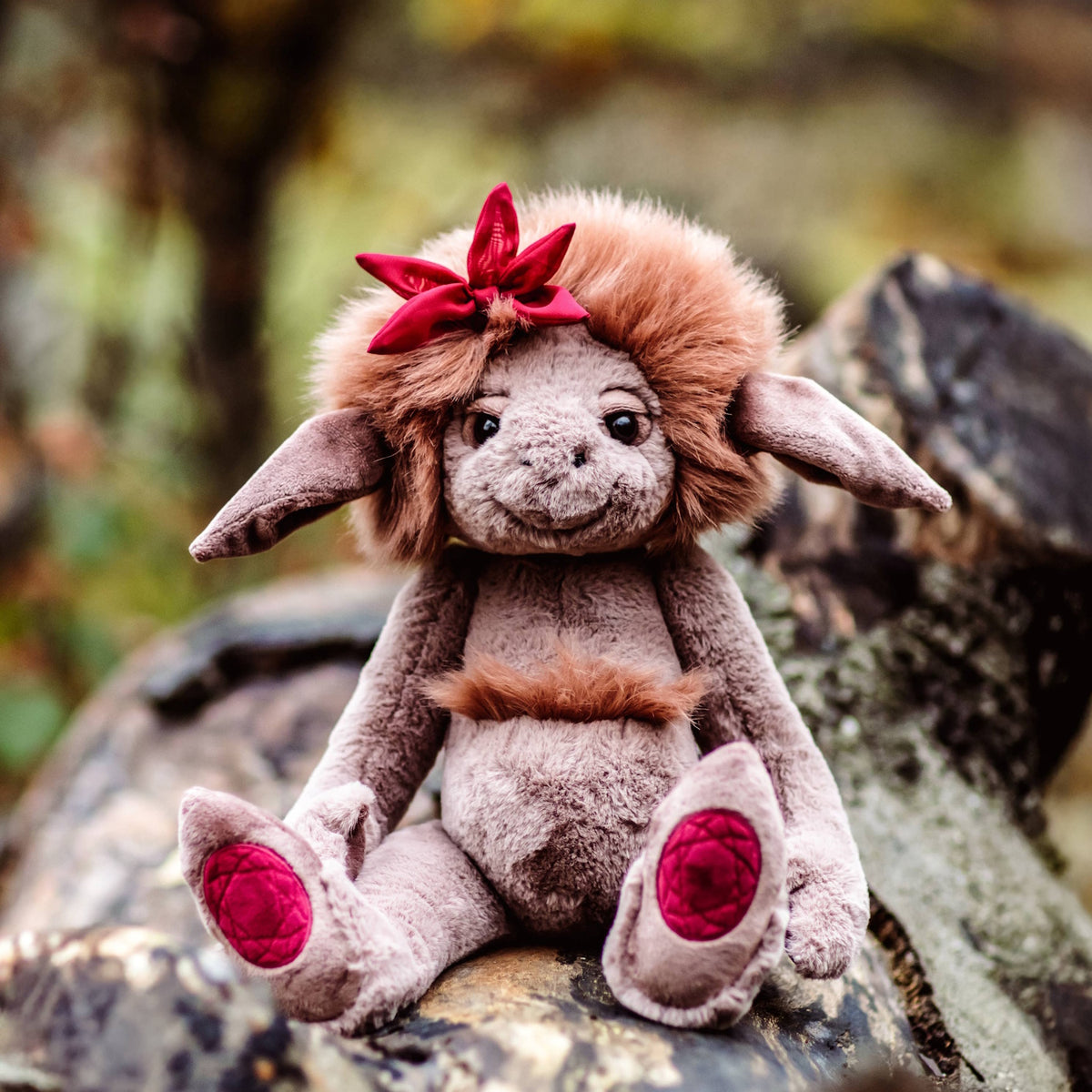 Plush creature with grey fur, brown mane, and red bow sitting outdoors on a wooden log in a natural setting.