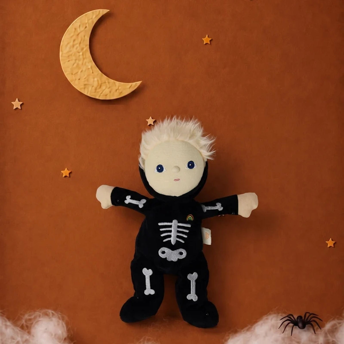 Skeleton plush doll set against a brown backdrop with a crescent moon, stars, cobwebs, and a spider.
