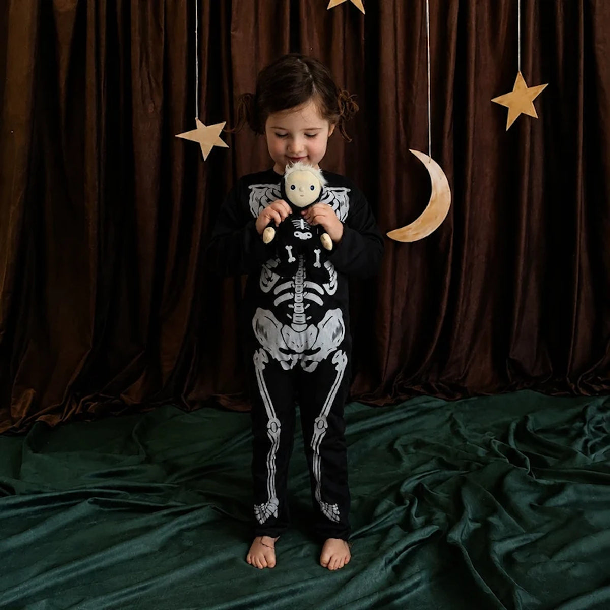 Young child in a skeleton outfit holding a matching skeleton plush doll against dark curtains.