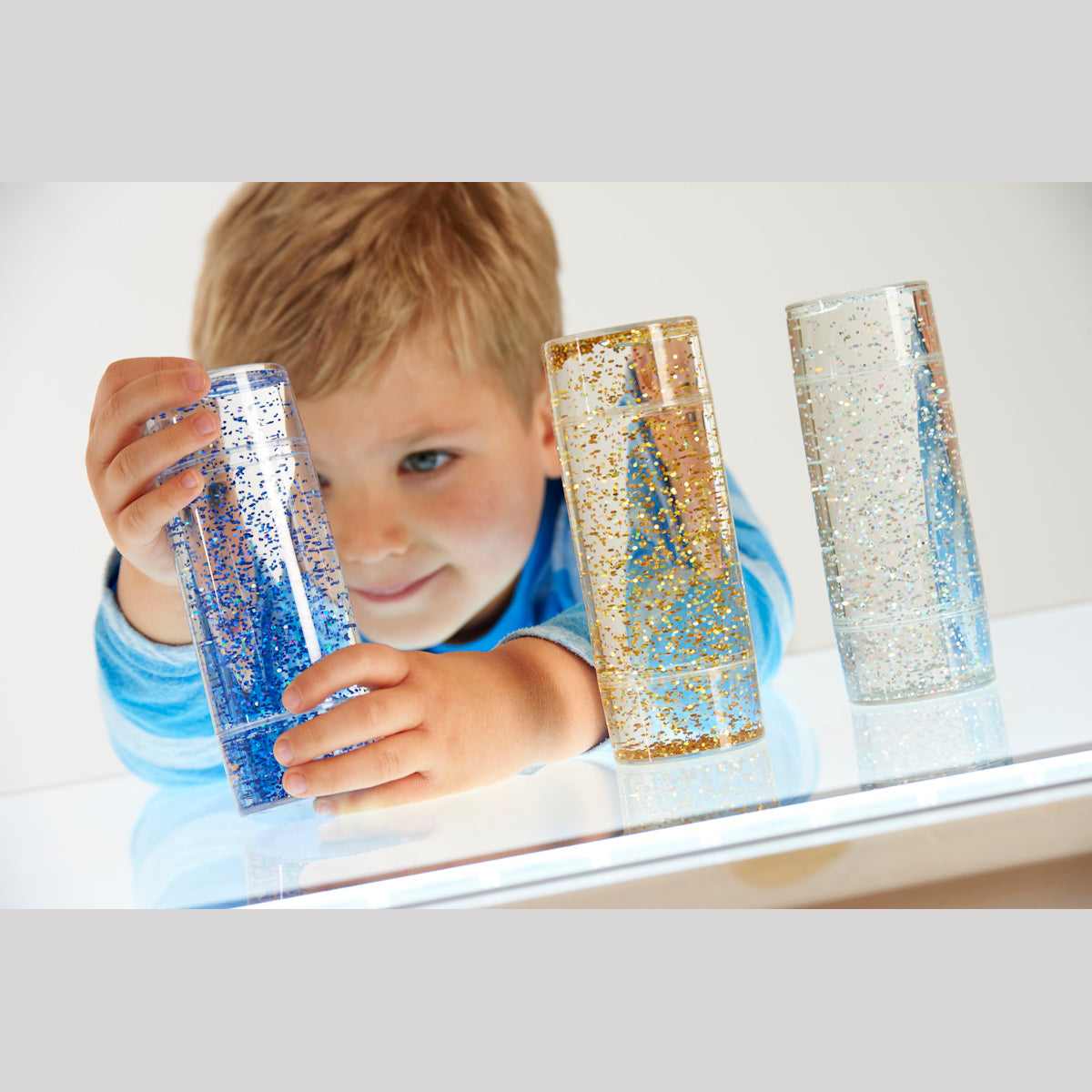 A child in a blue striped shirt is smiling and holding a clear cylinder filled with blue glitter. Two similar glitter cylinders are on a white table.