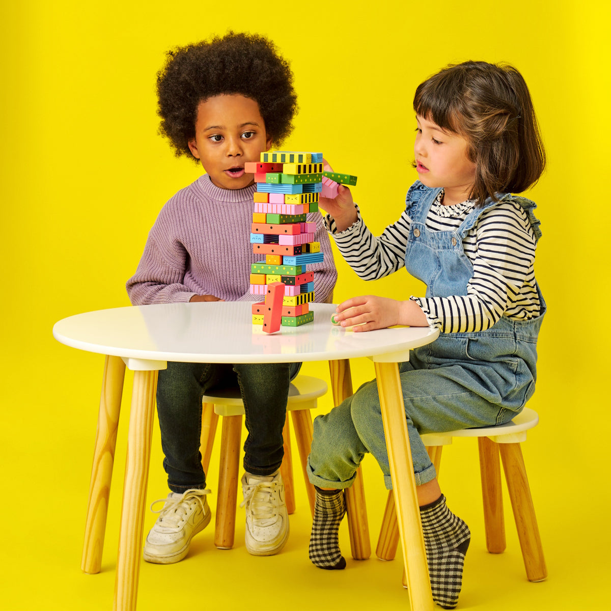 Two children focused on carefully stacking colorful wooden blocks on a white table against a yellow backdrop.