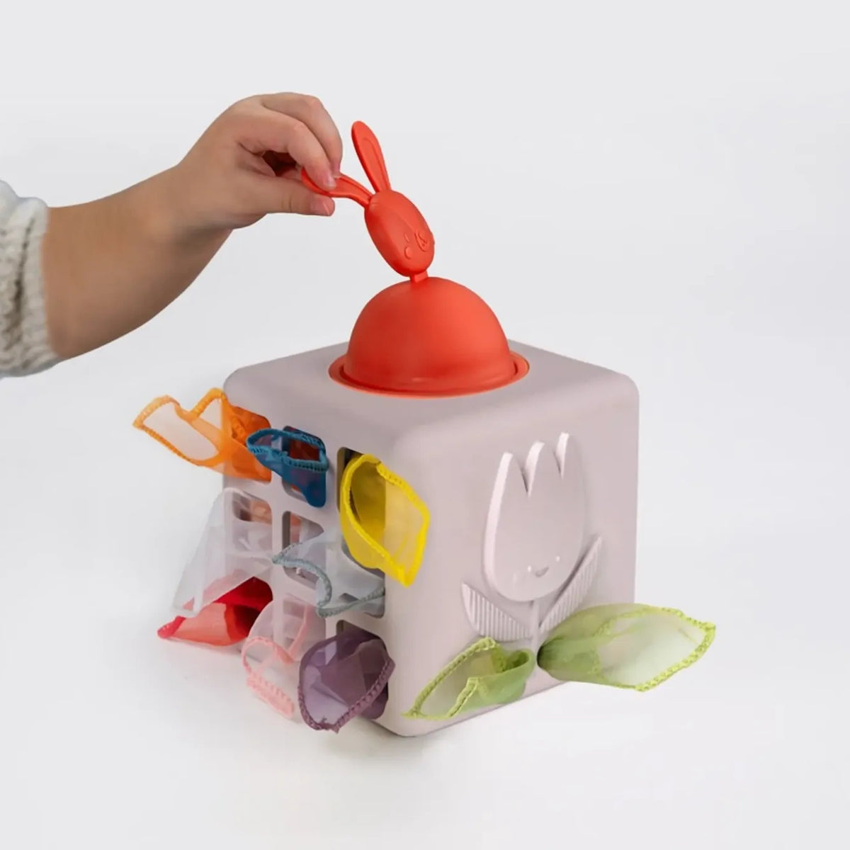 Child's hand pulling a red bunny topper on a sensory tissue cube filled with colorful fabric scarves.