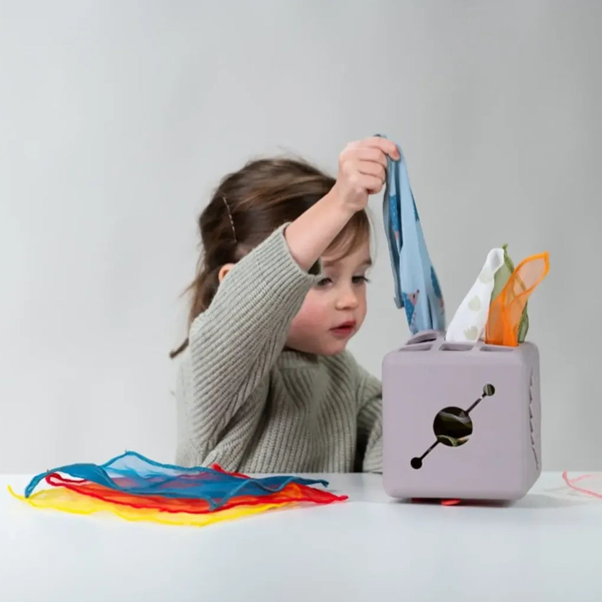 Toddler playing with a grey activity cube toy, pulling colorful fabric scarves out from top slots.