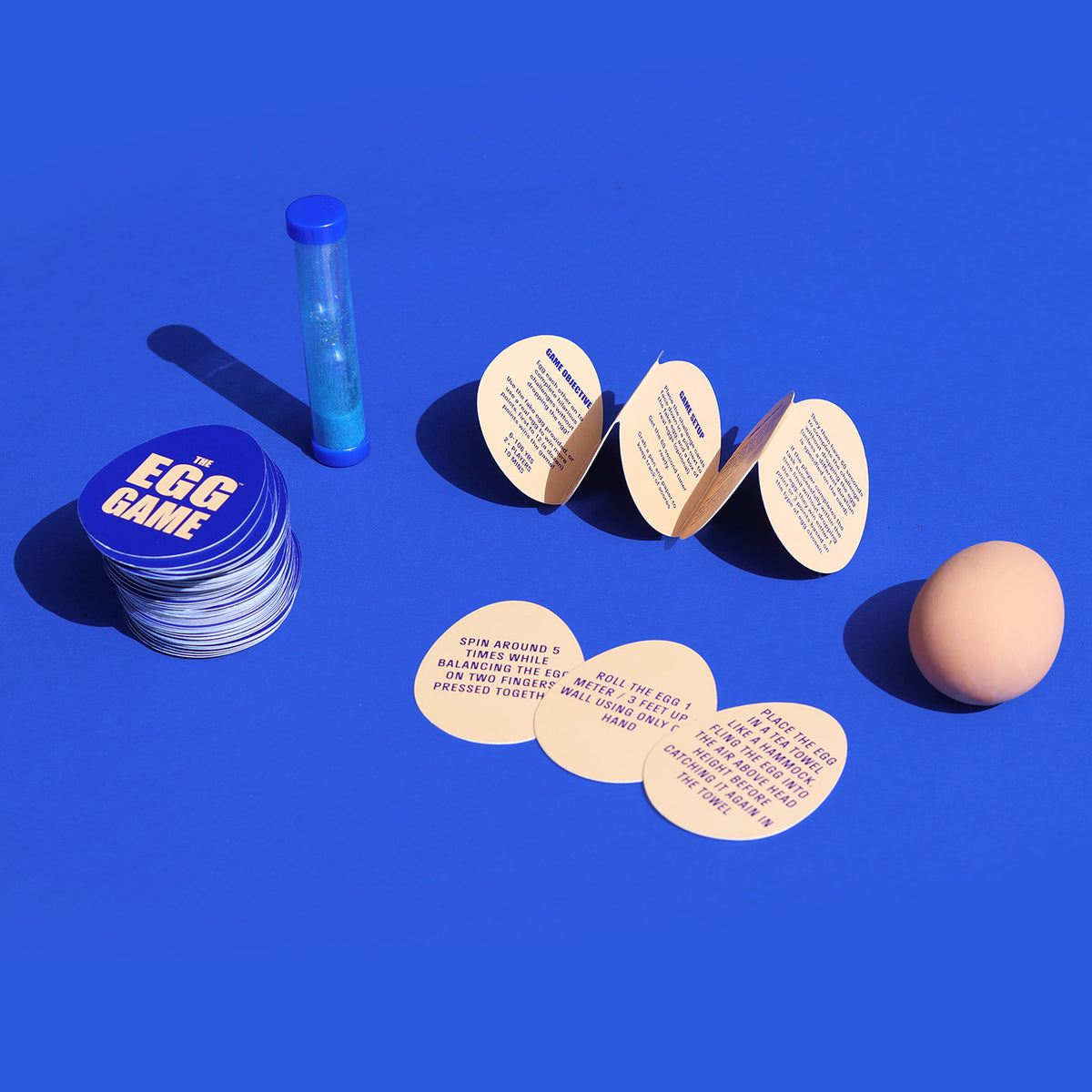 The Egg Game set with stack of egg cards, instruction accordion, sand timer, and egg model on blue surface.
