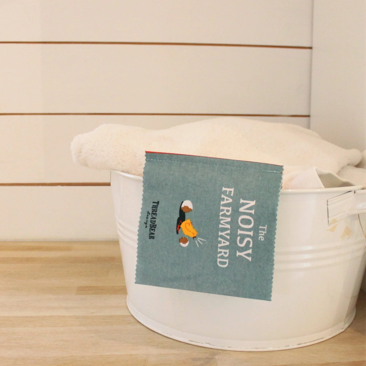 A fabric book cover titled "The Noisy Farmyard" hanging over the edge of a white metal tub.