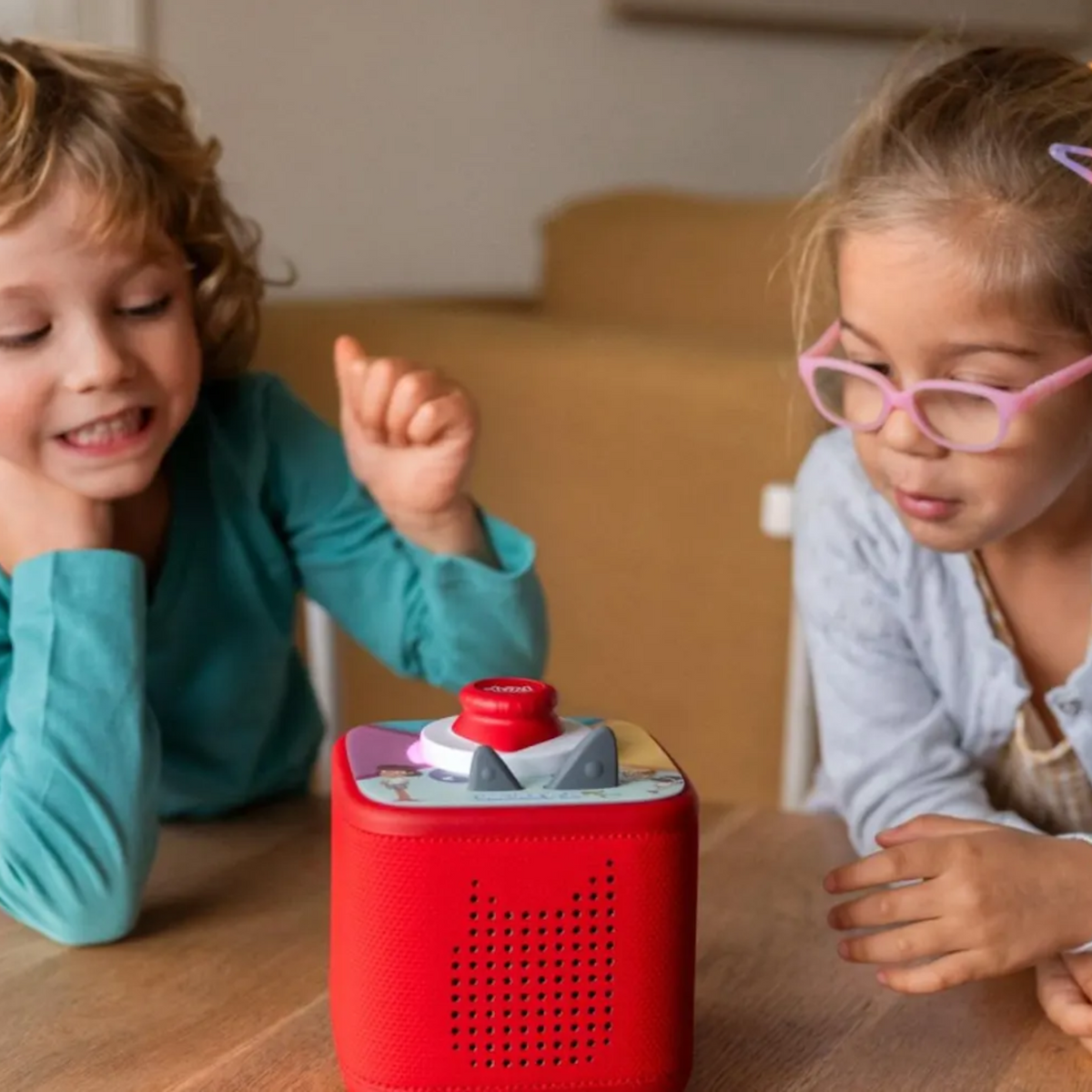 Two young children sitting at a wooden table, listening to a red Toniebox audio player.