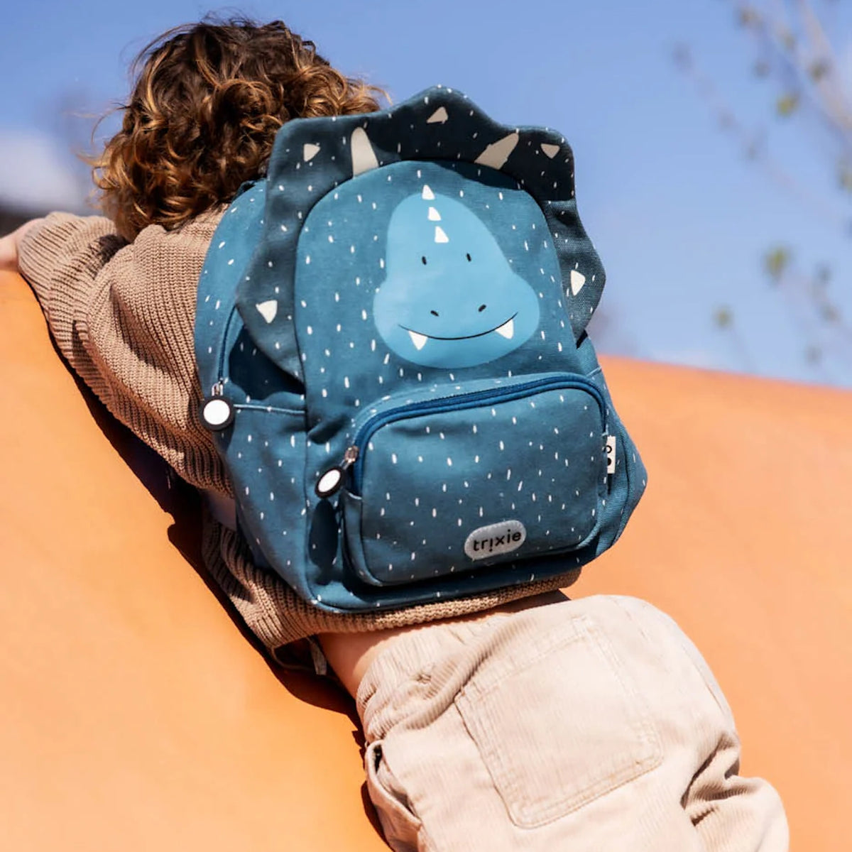 A child climbing a structure while wearing a blue triceratops dinosaur backpack with white speckles.