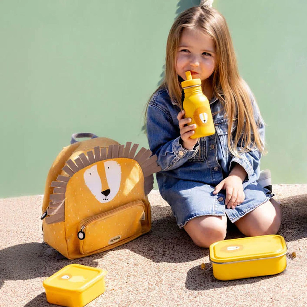 Young girl drinking from a yellow water bottle next to a matching lion backpack and lunch boxes.