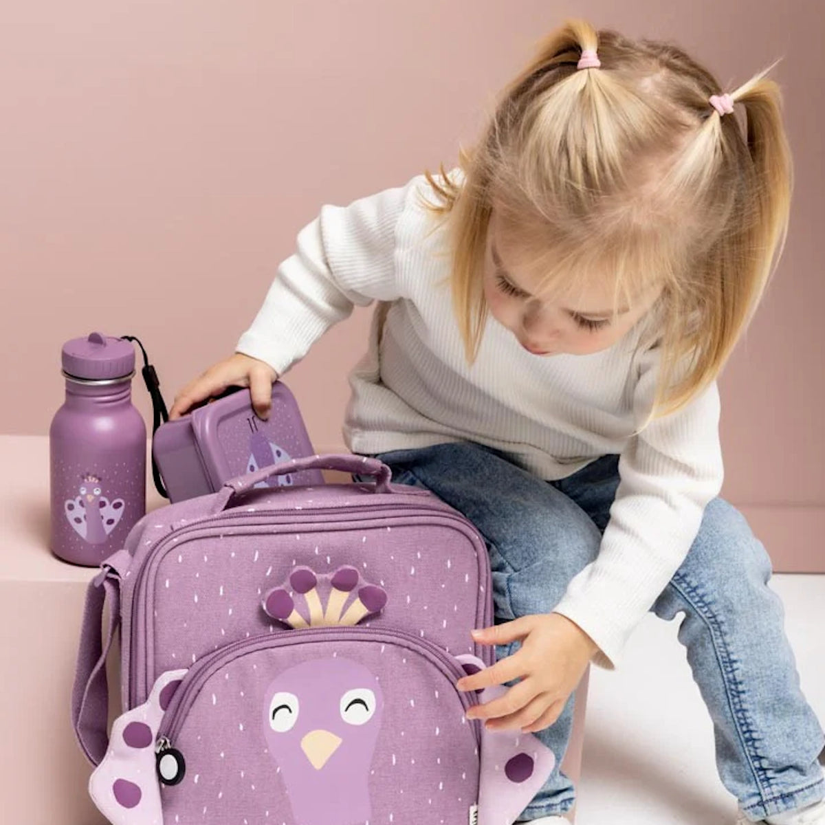 Young girl with pigtails reaching for a matching purple peacock bag, water bottle, and lunchbox.