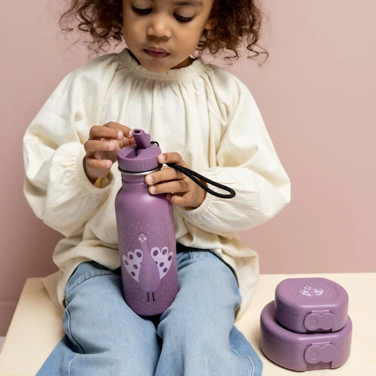Young girl holding a purple peacock water bottle next to matching snack containers against a pink backdrop.