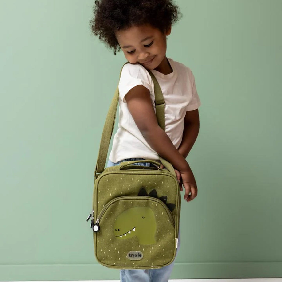 Young child carrying a green dinosaur-themed lunch bag over their shoulder against a solid green background.