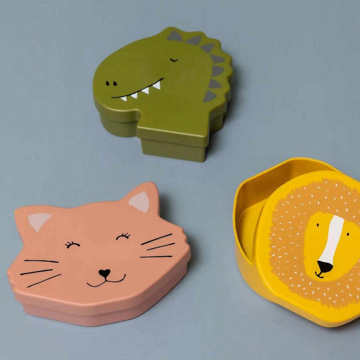 Three animal-shaped kids' lunch boxes in green dinosaur, pink cat, and yellow lion designs on a gray background.
