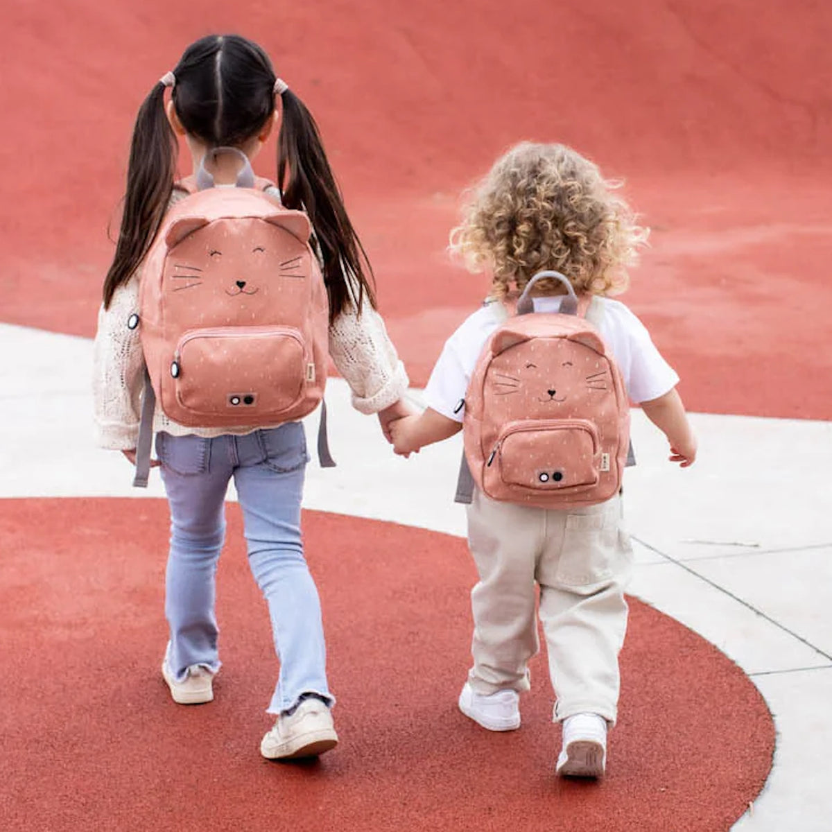 Two children seen from behind holding hands, each wearing a pink cat-themed backpack on a playground.