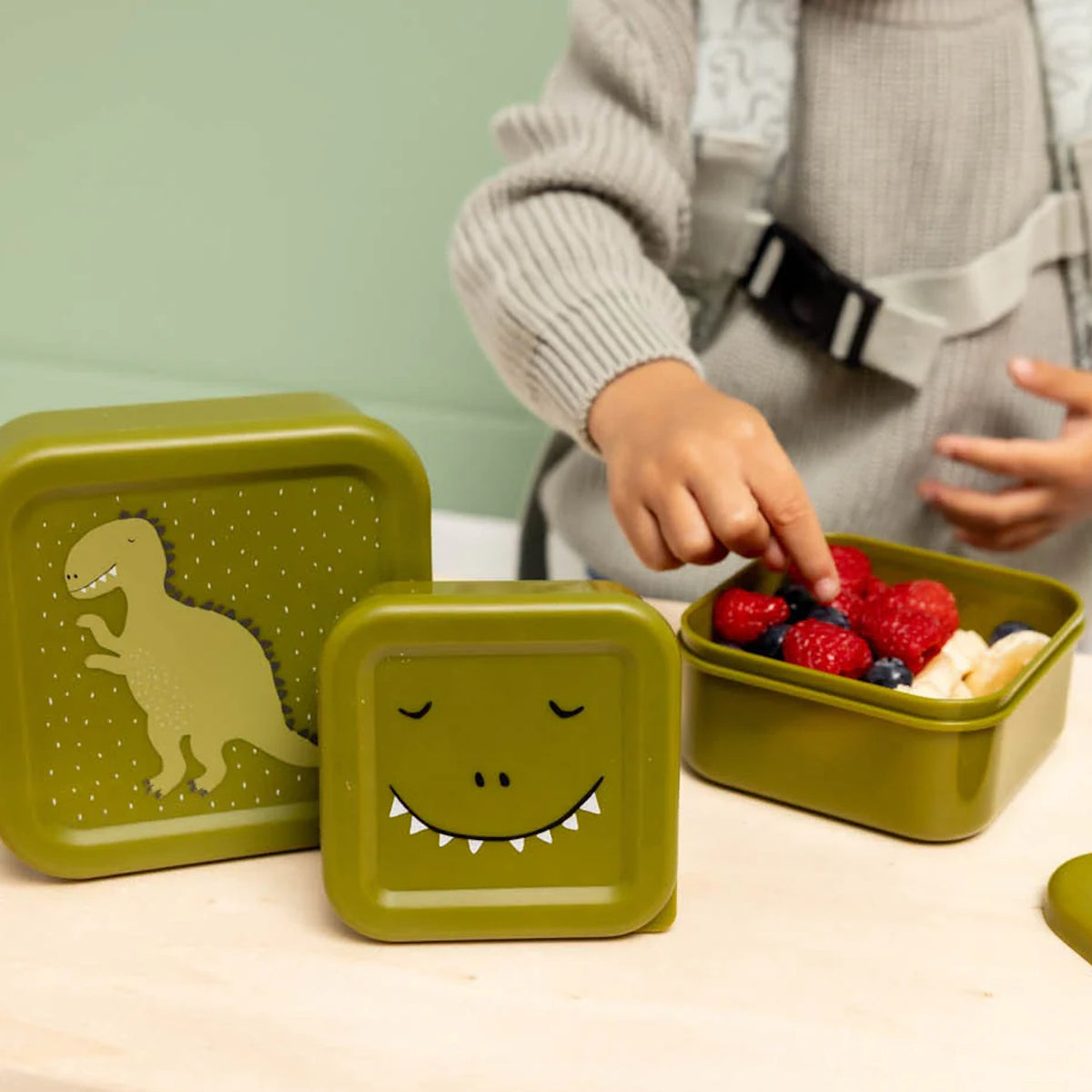 A child reaches into an open green dinosaur snack box filled with fresh berries and banana slices.