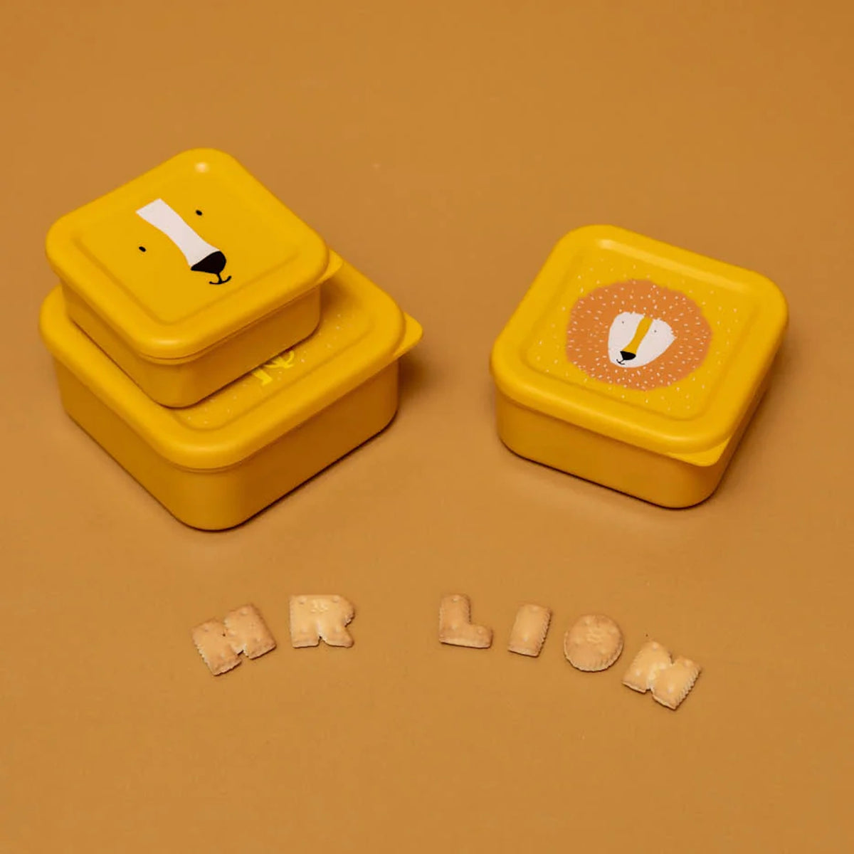 Three yellow lion-themed nesting snack boxes displayed with alphabet crackers on a solid ochre background.