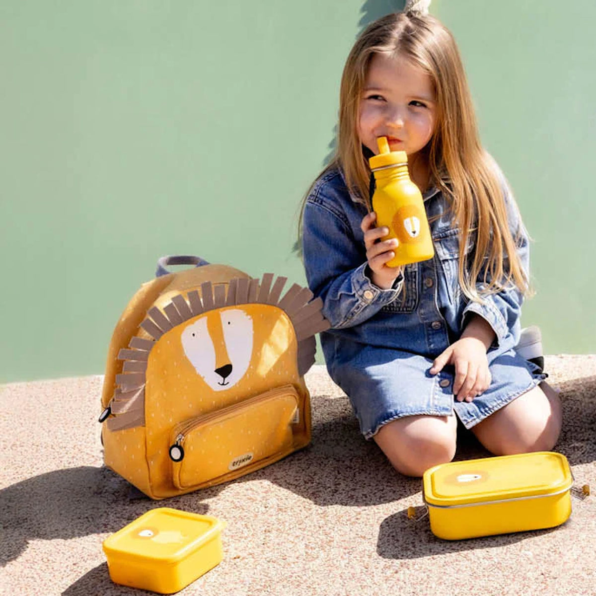 A young girl in a denim dress sits outside with a yellow lion-themed backpack, water bottle, and lunch boxes.